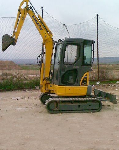 Miniexcavadora amarilla en un terreno baldío cerca de una valla de malla, con el brazo levantado.