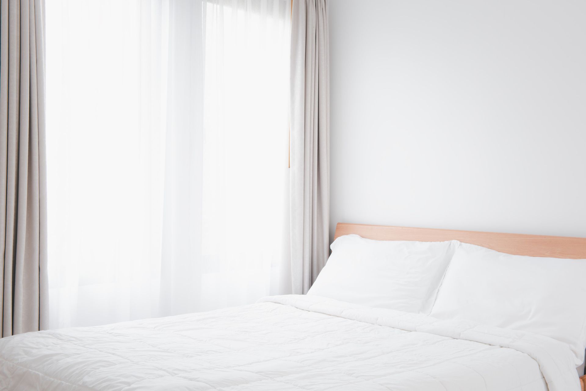 Una cama con sábanas blancas y almohadas en un dormitorio al lado de una ventana.
