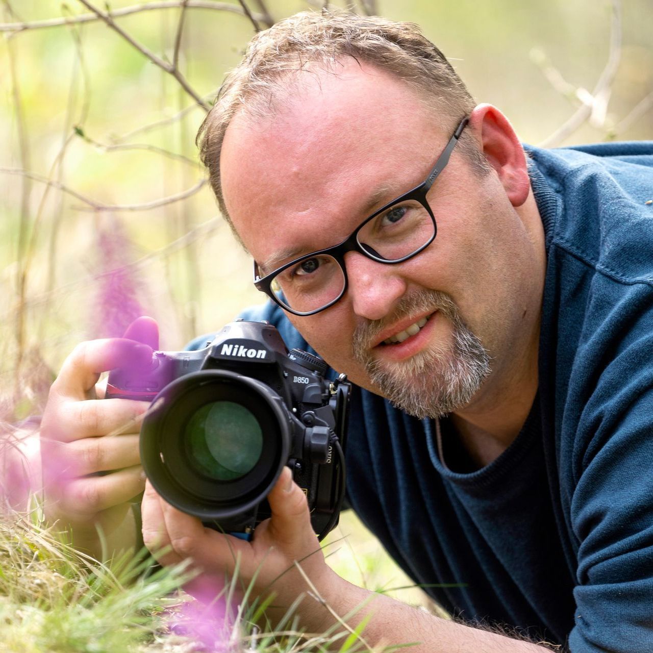 Mann mit Brille hält eine Nikon-Kamera und lächelt, während er im Freien ein Foto macht.