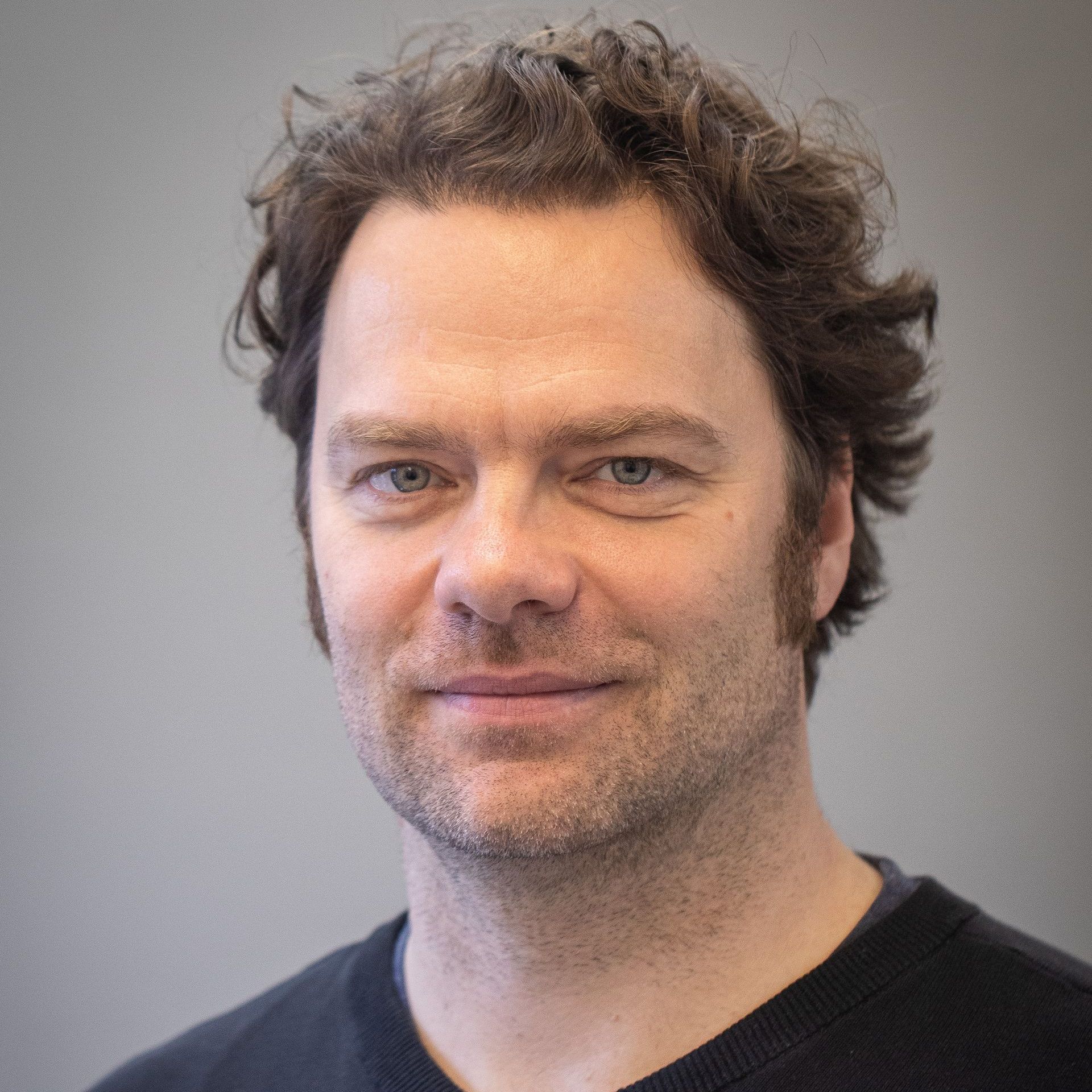 Lächelnder Mann mit lockigem braunem Haar, schwarzes Hemd, vor einer grauen Wand.