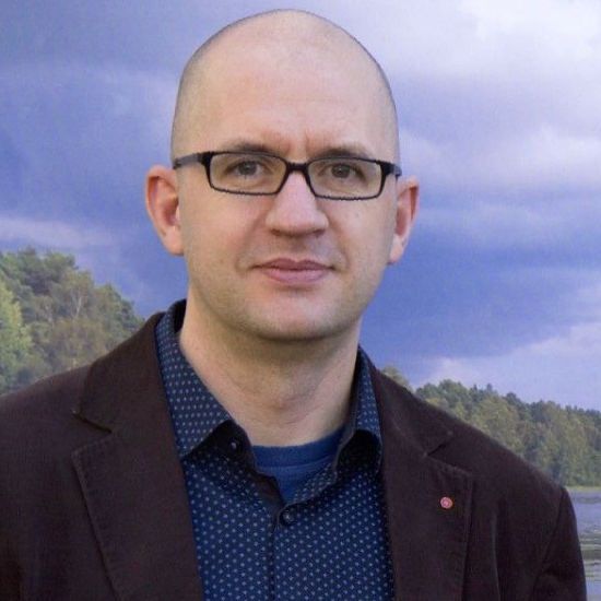 Glatzköpfiger Mann mit Brille, brauner Jacke und blau gemustertem Hemd steht vor einer verschwommenen Szene im Freien.