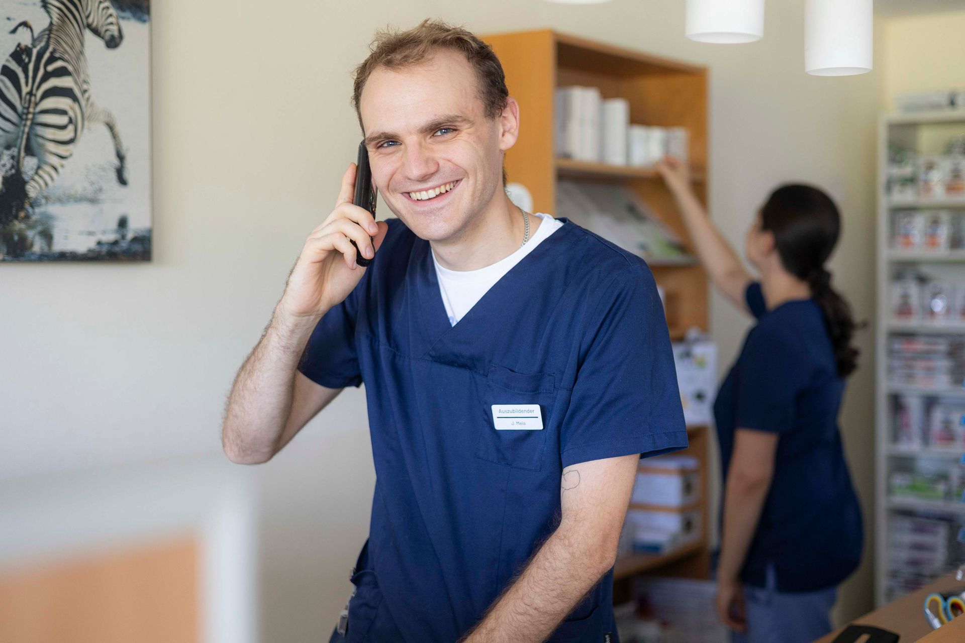 Ein lächelnder Mann in blauer OP-Kleidung telefoniert in einem hellen Empfangsbereich einer Klinik.