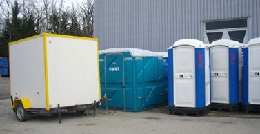 Une remorque utilitaire blanche, une grande unité portable turquoise et trois toilettes portables bleues et blanches sont stationnées près d'un bâtiment gris.