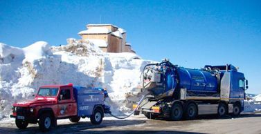 Un pick-up rouge et un camion-citerne bleu sont garés sur une route, à côté d'un gros tas de neige et d'un bâtiment.