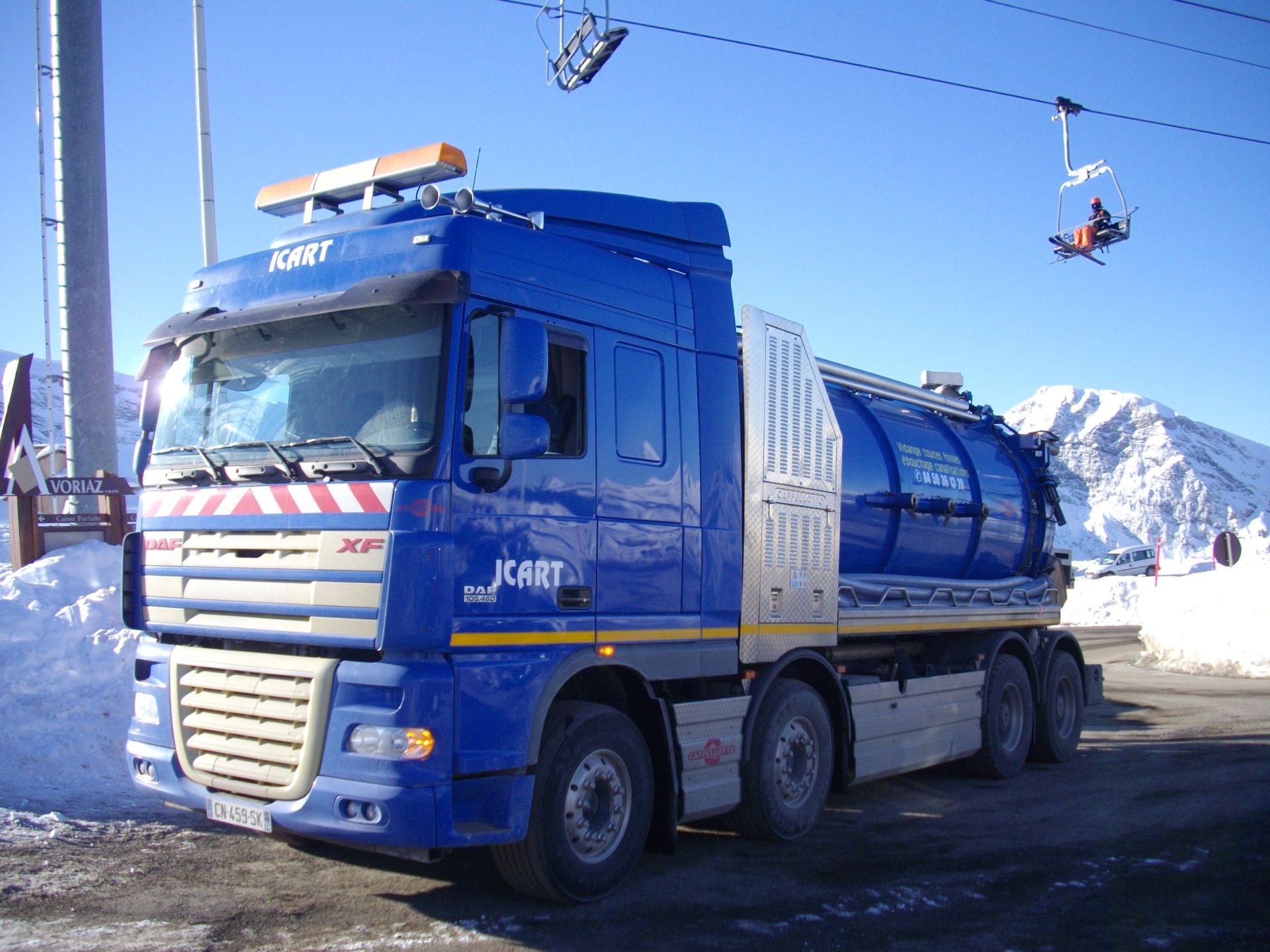 Un camion-citerne Jcart bleu est stationné sur une route de montagne enneigée, avec un télésiège visible au-dessus.
