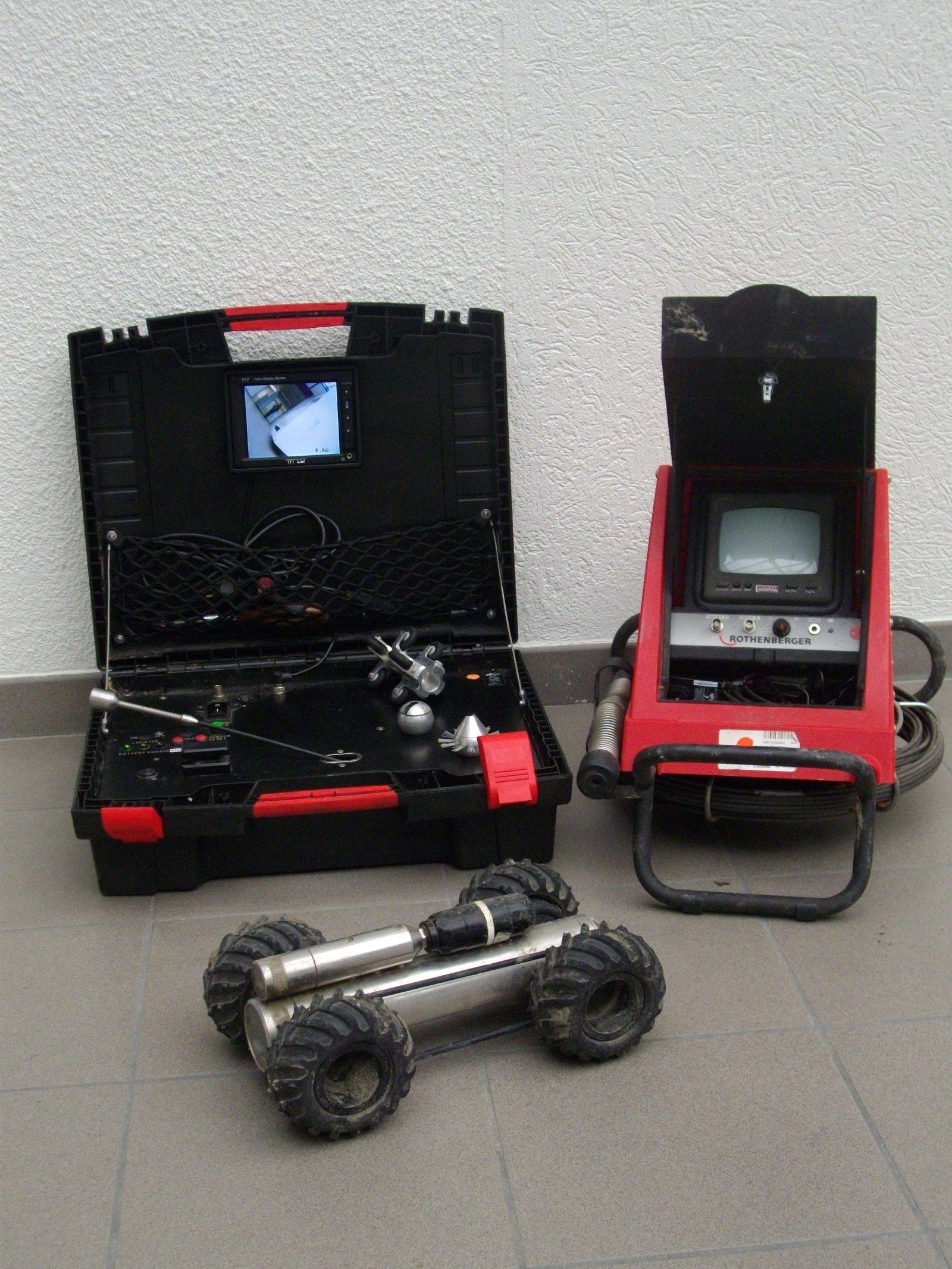 Un robot d'inspection de canalisations portable, équipé d'une caméra, d'un boîtier noir et d'une unité de commande rouge avec un écran, est posé sur un sol carrelé.