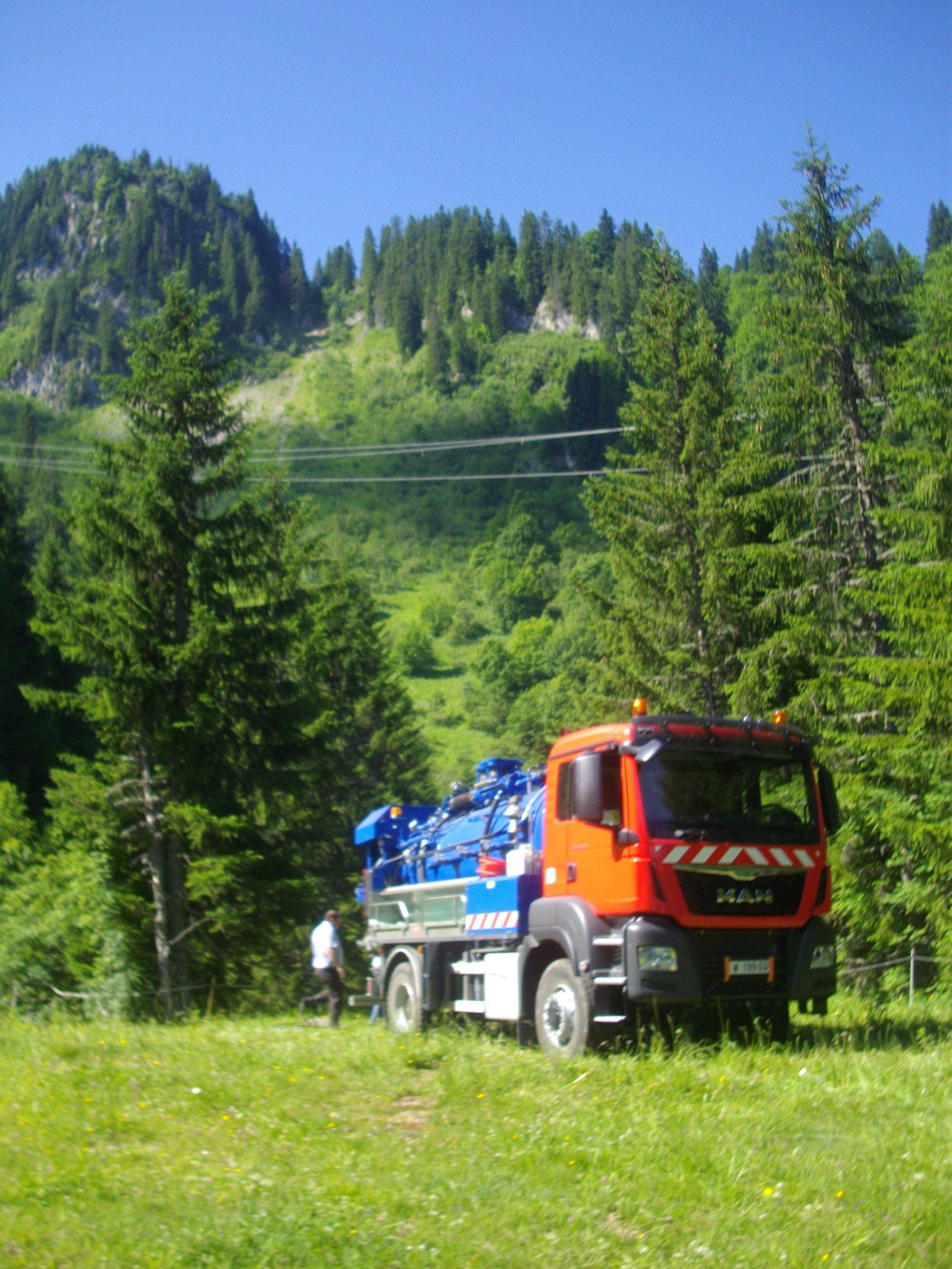 Un camion-citerne bleu et orange est stationné sur le flanc d'une colline montagneuse herbeuse, avec une forêt dense en arrière-plan, sous un ciel dégagé.
