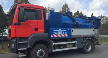 Un camion aspirateur industriel rouge et bleu est garé à l'extérieur, sur fond d'arbres et de ciel nuageux.