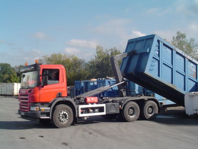 Un camion Scania orange à bras de levage charge un grand conteneur rectangulaire bleu dans une cour extérieure.
