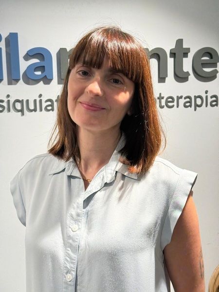 Una mujer con cabello castaño ondulado, ojos azules y lápiz labial rosa sonríe, apoyando la mano en su mejilla.