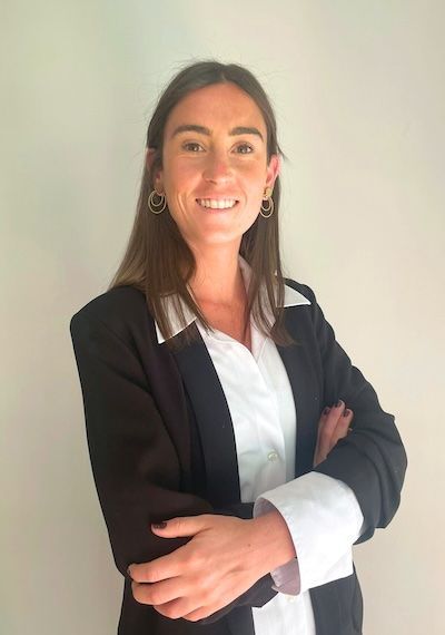 Una mujer con cabello castaño ondulado, ojos azules y lápiz labial rosa sonríe, apoyando la mano en su mejilla.