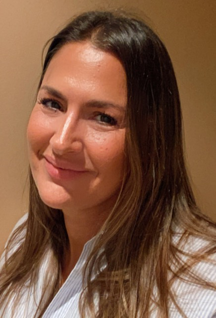 Una mujer con cabello castaño ondulado, ojos azules y lápiz labial rosa sonríe, apoyando la mano en su mejilla.