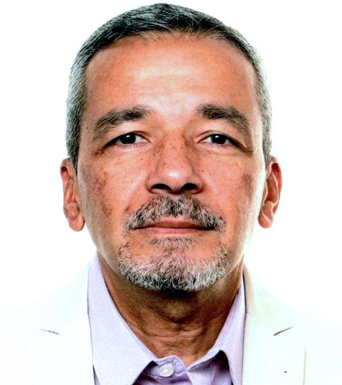 Hombre de pelo canoso y perilla, vestido con chaqueta blanca y camisa morada, mirando a la cámara.