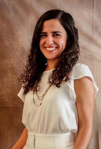 Una mujer con cabello castaño ondulado, ojos azules y lápiz labial rosa sonríe, apoyando la mano en su mejilla.