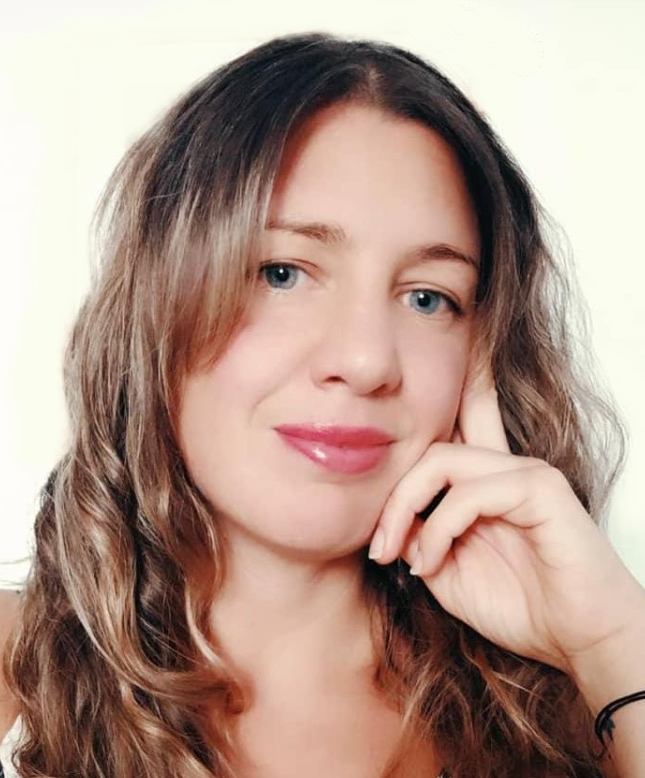 Una mujer con cabello castaño ondulado, ojos azules y lápiz labial rosa sonríe, apoyando la mano en su mejilla.