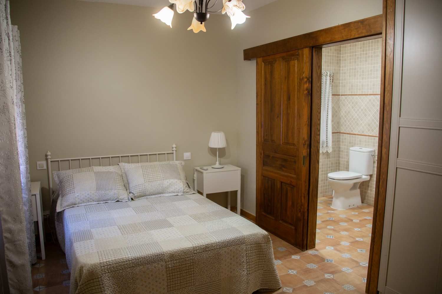 Habitación pequeña con cama blanca, mesita de noche y puerta corredera que da al baño con inodoro.