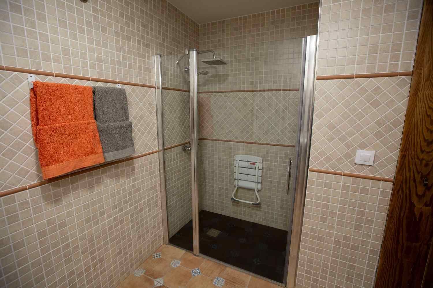Cabina de ducha alicatada con puertas de cristal, toallero y toallas naranjas y grises colgadas en la pared.
