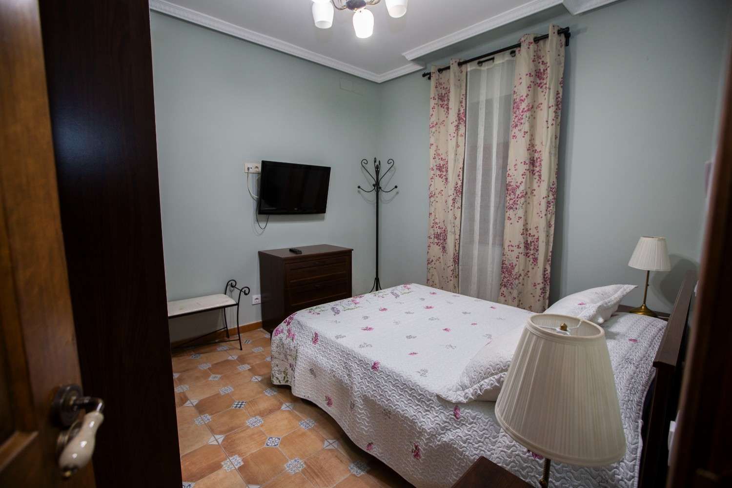 Acogedora habitación con cama, televisor montado en la pared, cómoda, lámpara y cortinas florales.