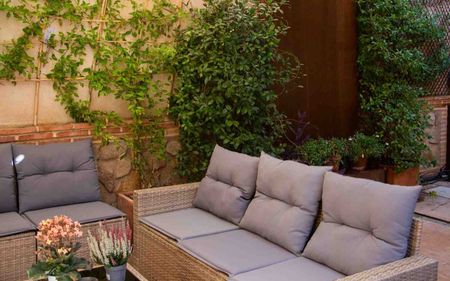 Patio de piedra con mesa y sillas de madera bajo una pérgola junto a un jardín lleno de flores y un muro de estilo rústico.