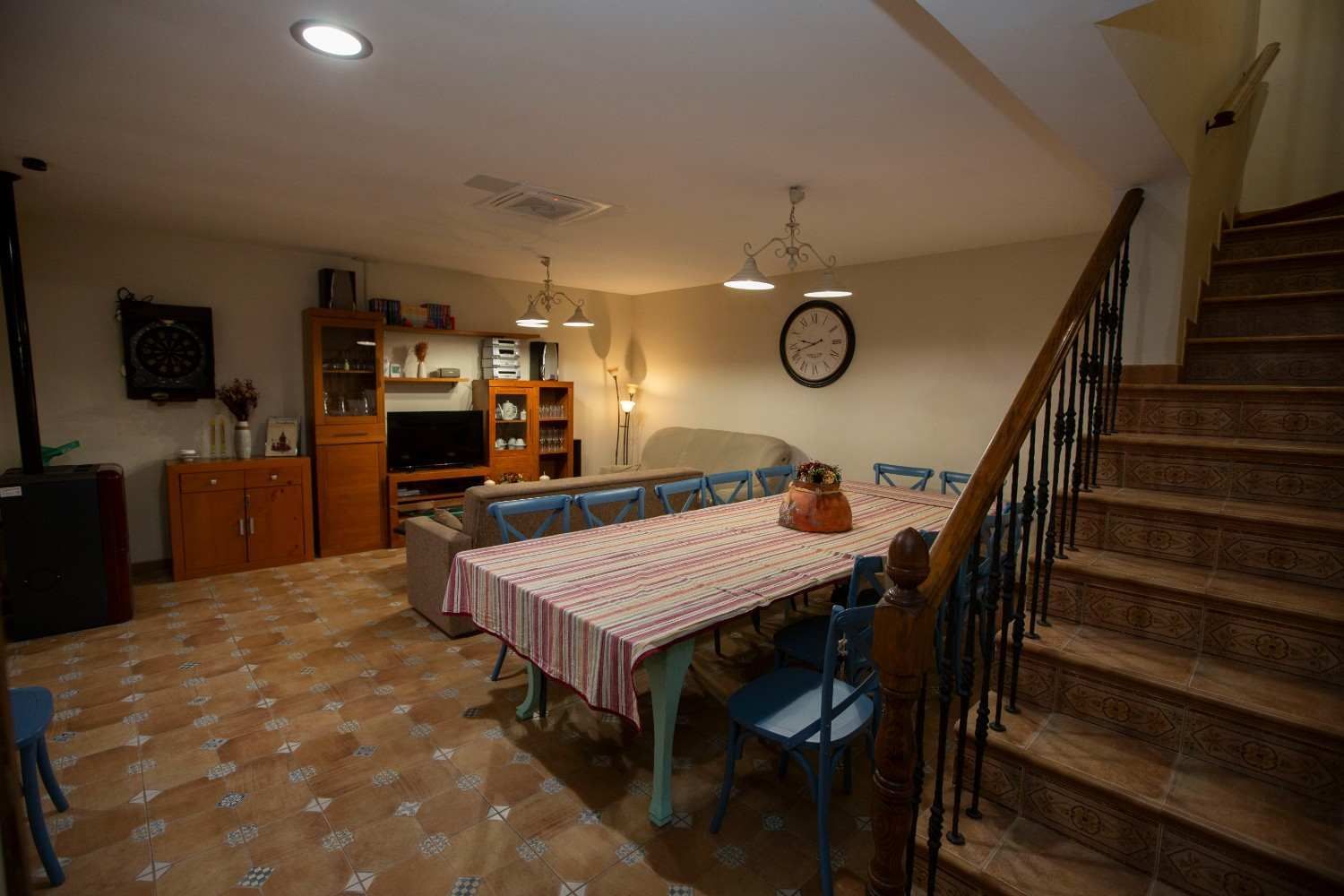 Comedor rústico con mesa de madera, bancos, plantas en macetas y luz solar que entra por dos ventanas.