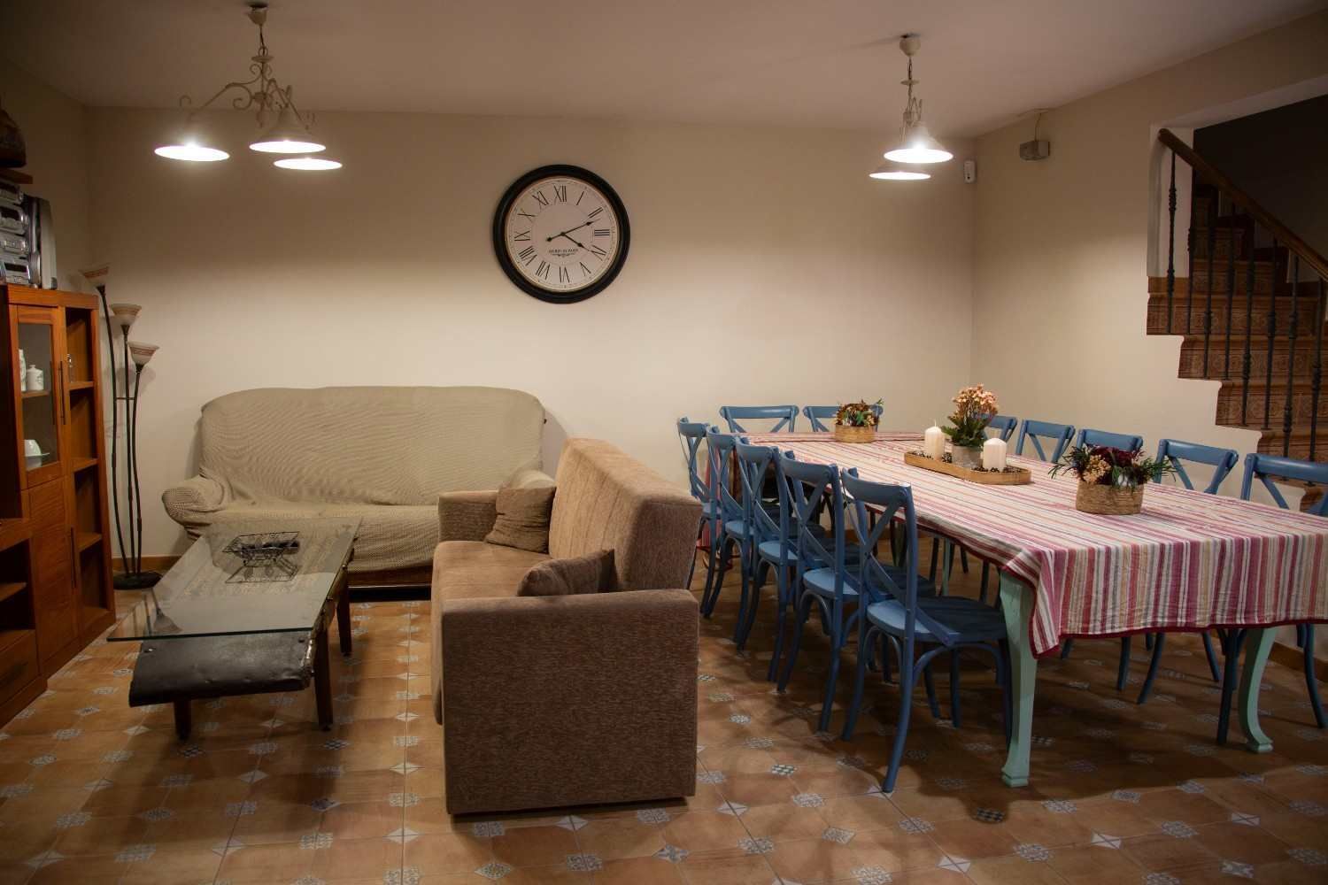 Comedor rústico con mesa de madera, bancos, plantas en macetas y luz solar que entra por dos ventanas.