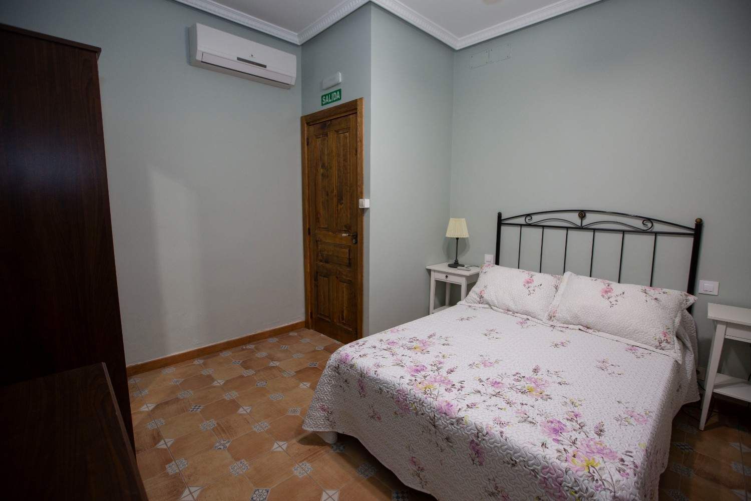 Habitación pequeña con colcha floral, puerta de madera y suelo de baldosas.