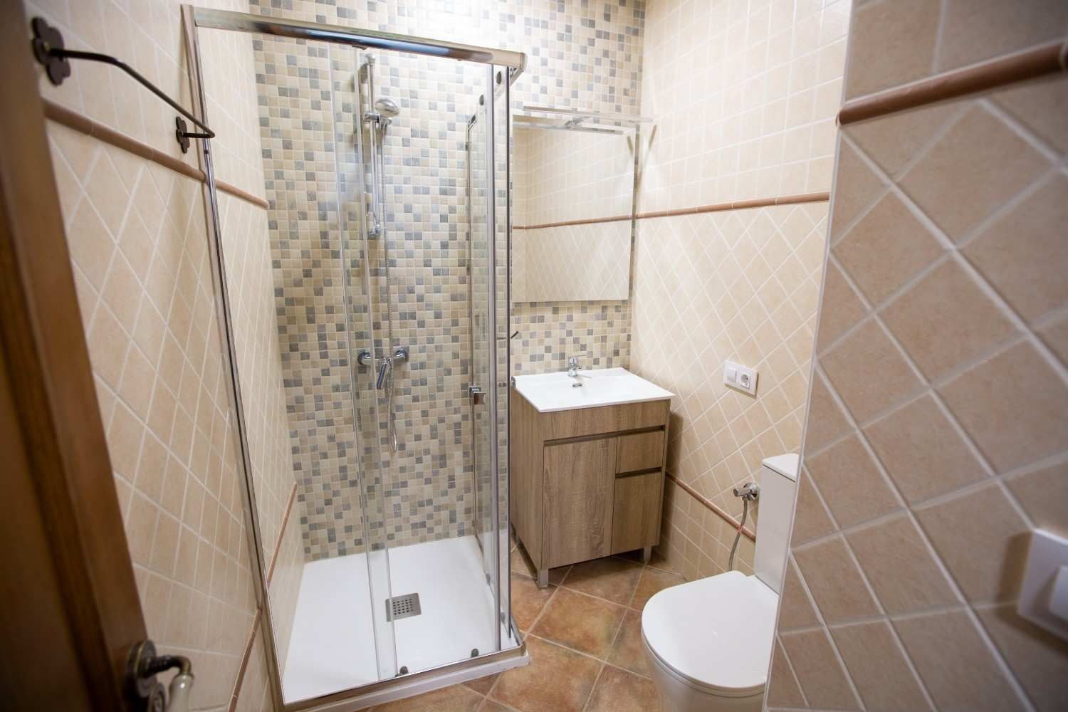 Pequeño baño de color beige con ducha, lavabo y inodoro suspendido, visto a través de una mampara de ducha de cristal.