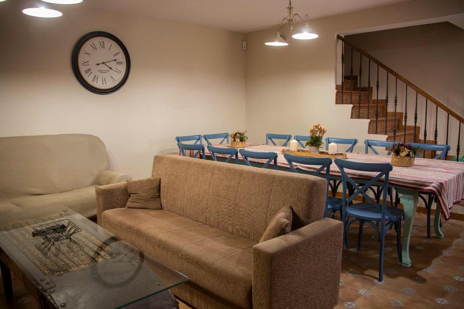 Comedor rústico con mesa de madera, bancos, plantas en macetas y luz solar que entra por dos ventanas.