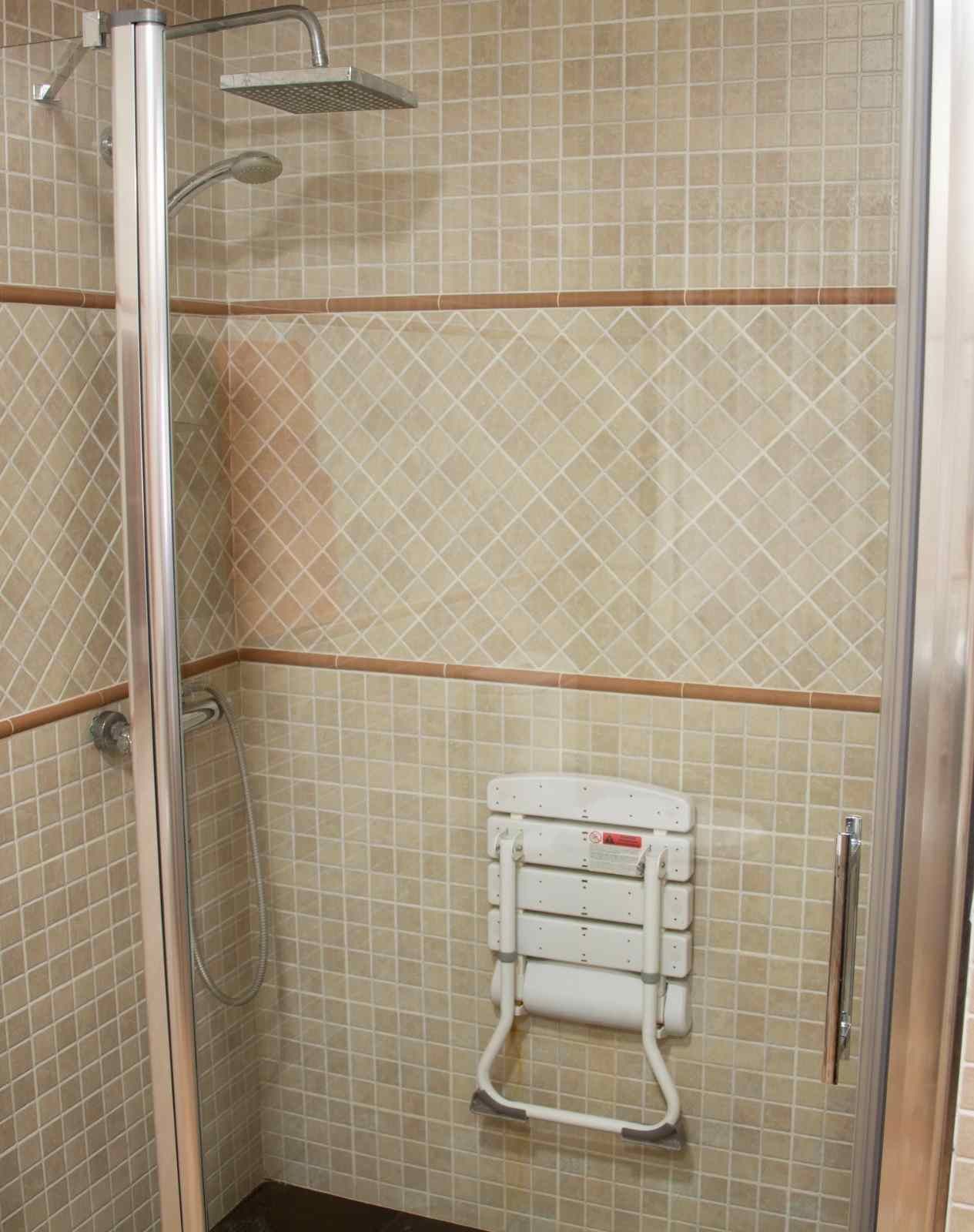Cabina de ducha con paredes alicatadas en color beige, puertas de cristal y un asiento plegable blanco montado en la pared.