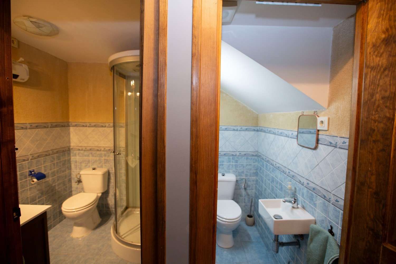 Vista dividida de dos pequeños baños con inodoros, lavabos y paredes alicatadas en tonos cálidos beige y azul.