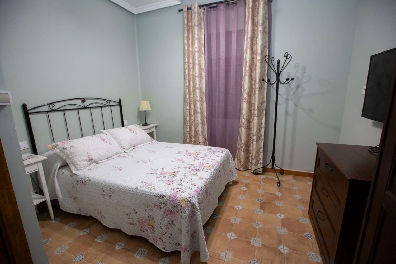 Dormitorio con cama, ropa de cama con estampado floral, cortinas moradas y una cómoda en una habitación pequeña.