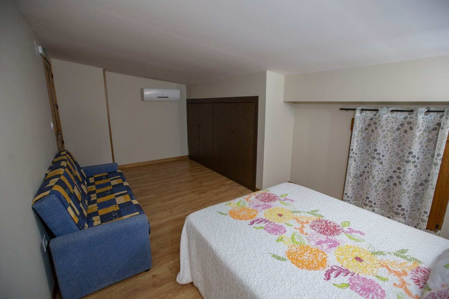 Habitación pequeña con sofá azul, colcha floral, suelo de madera, armario y ventana con cortinas.