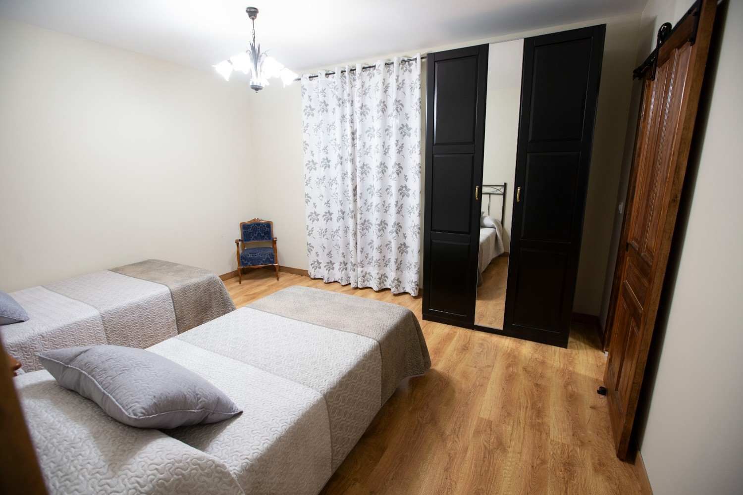 Habitación ordenada con dos camas grises, suelo de madera y un armario junto a una ventana con cortinas transparentes.