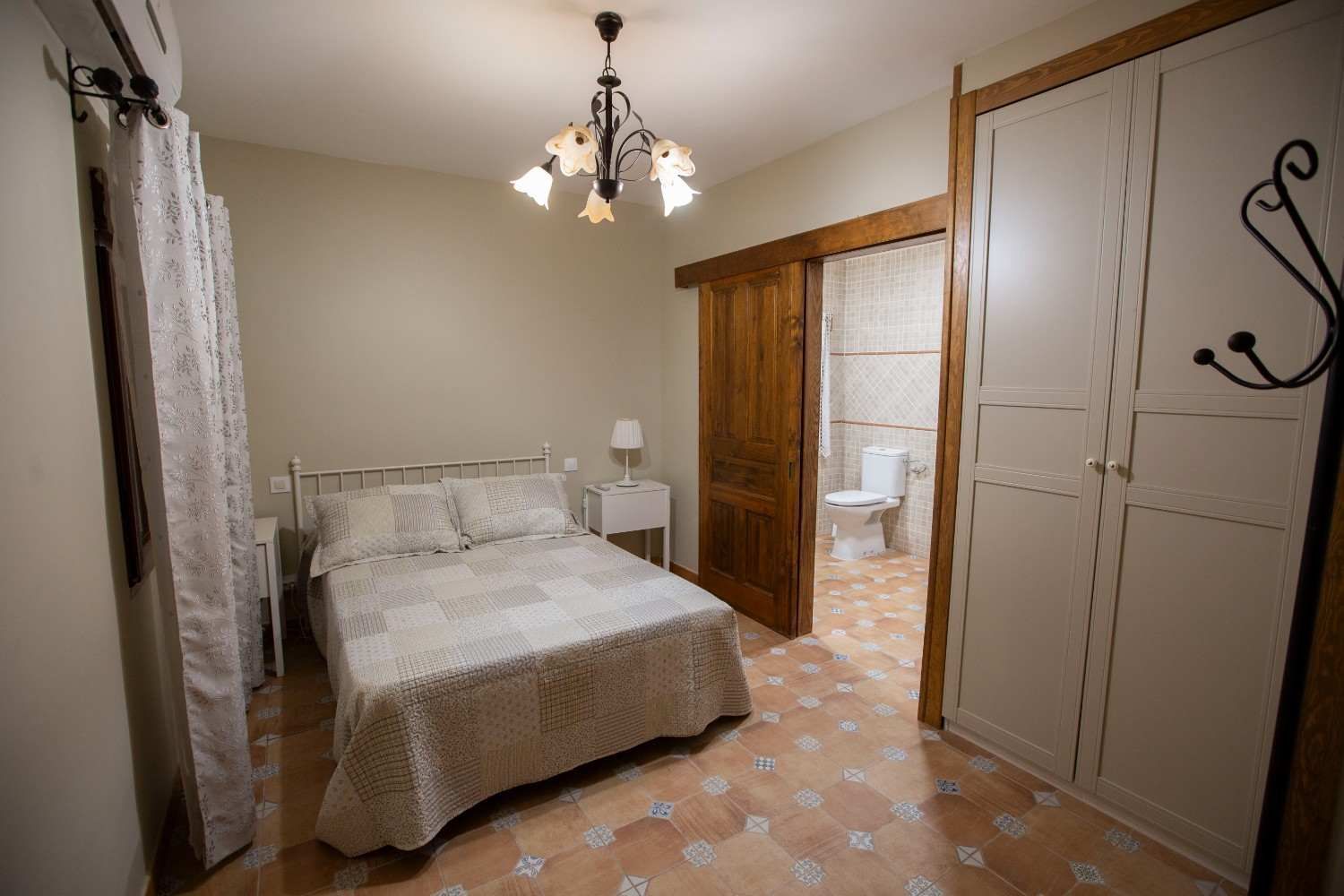 Dormitorio con cama, lámpara de araña, suelo de baldosas estampadas, puertas de armario y puerta abierta al baño.