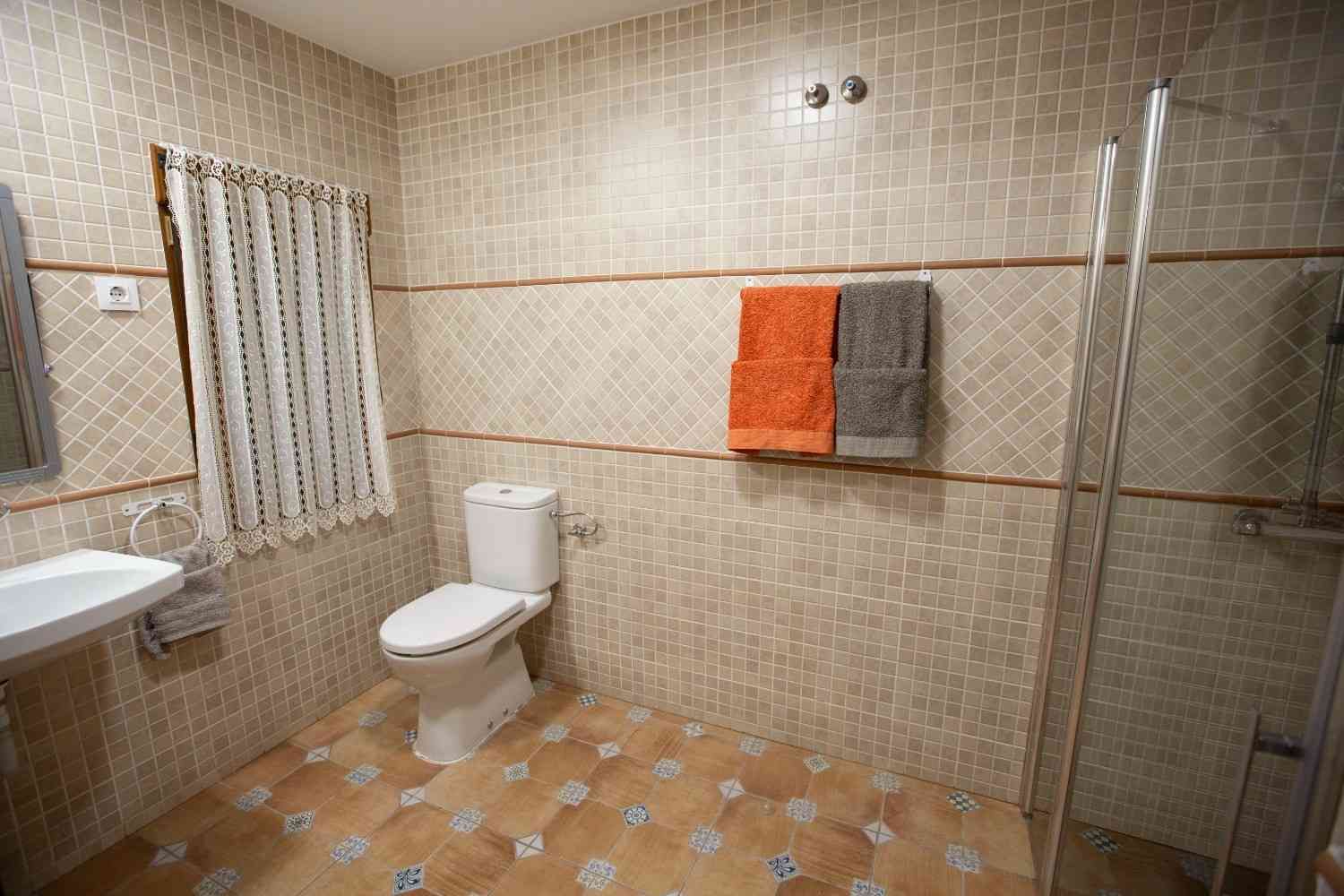Amplio baño con azulejos beige, inodoro, cabina de ducha, lavabo y toallero naranja en la pared.