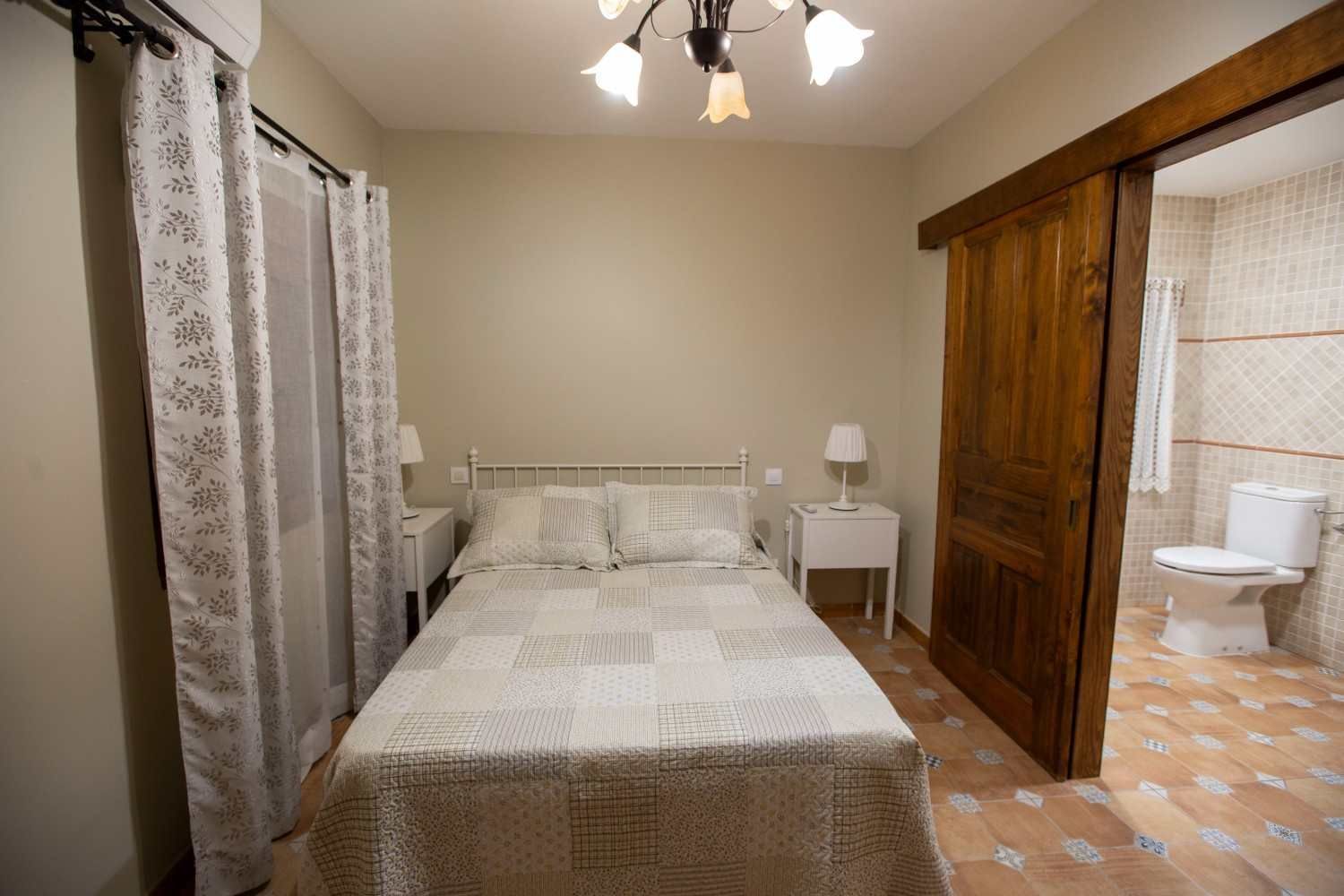 Acogedora habitación con dos camas individuales, cortinas de encaje y puerta abierta que da a un baño alicatado.