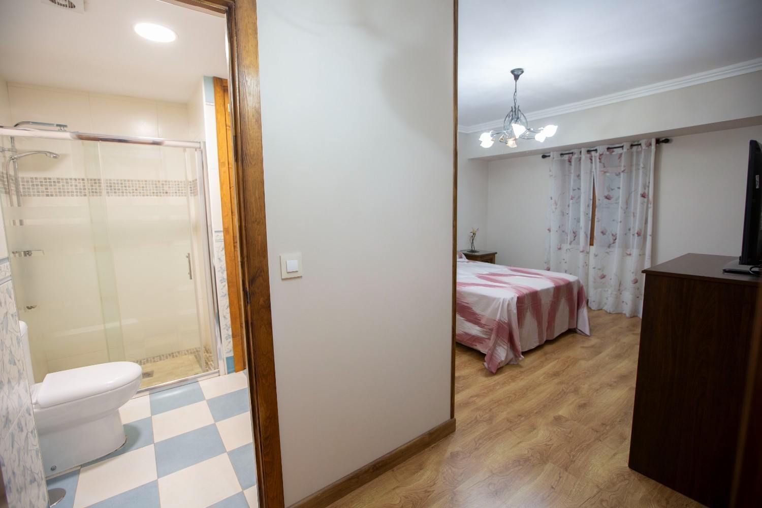 Habitación pequeña en apartamento con baño contiguo, suelo de madera, colcha rosa y zona de ducha alicatada.