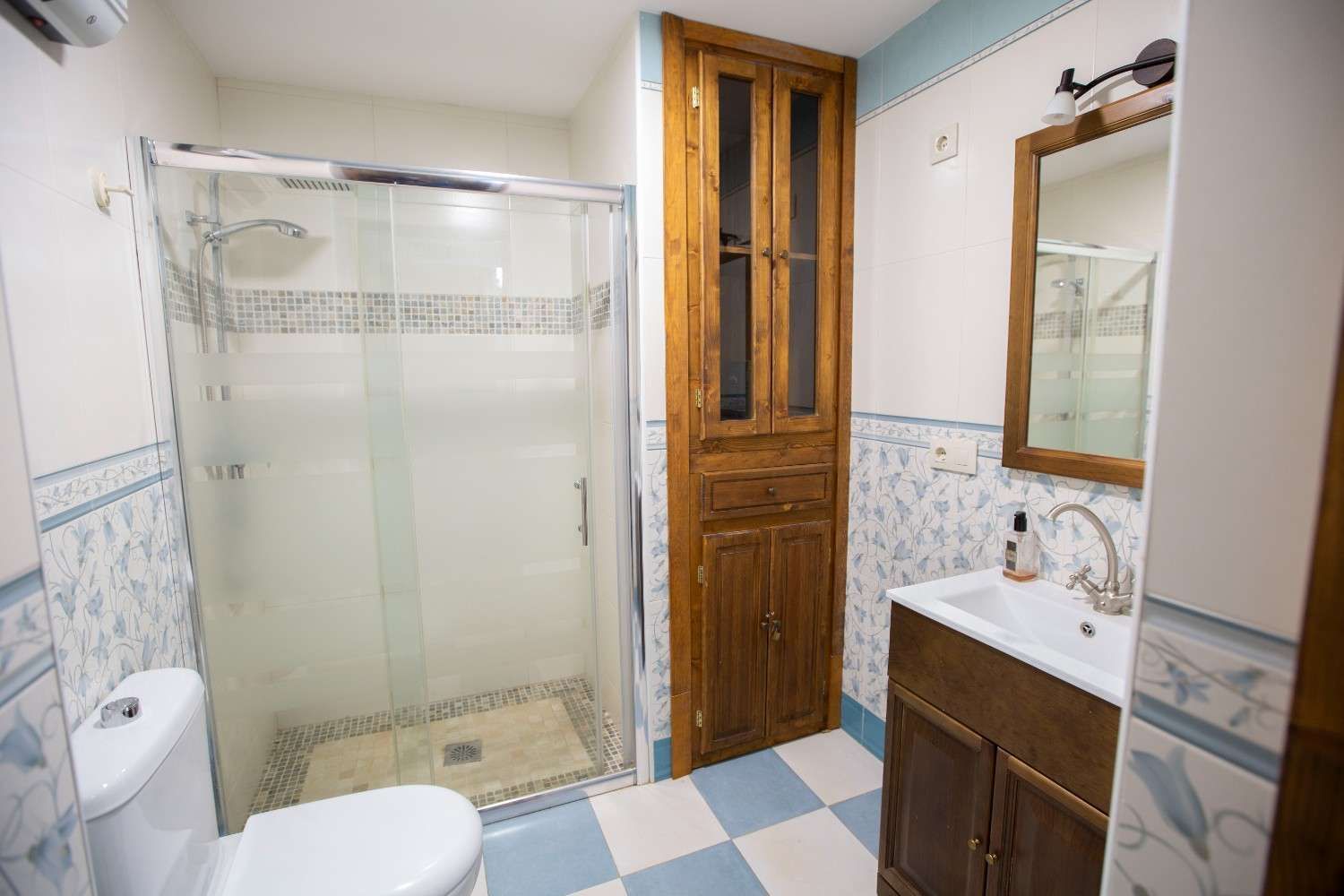 Baño luminoso con ducha de cristal, inodoro, lavabo, espejo y suelo de baldosas azules y blancas.