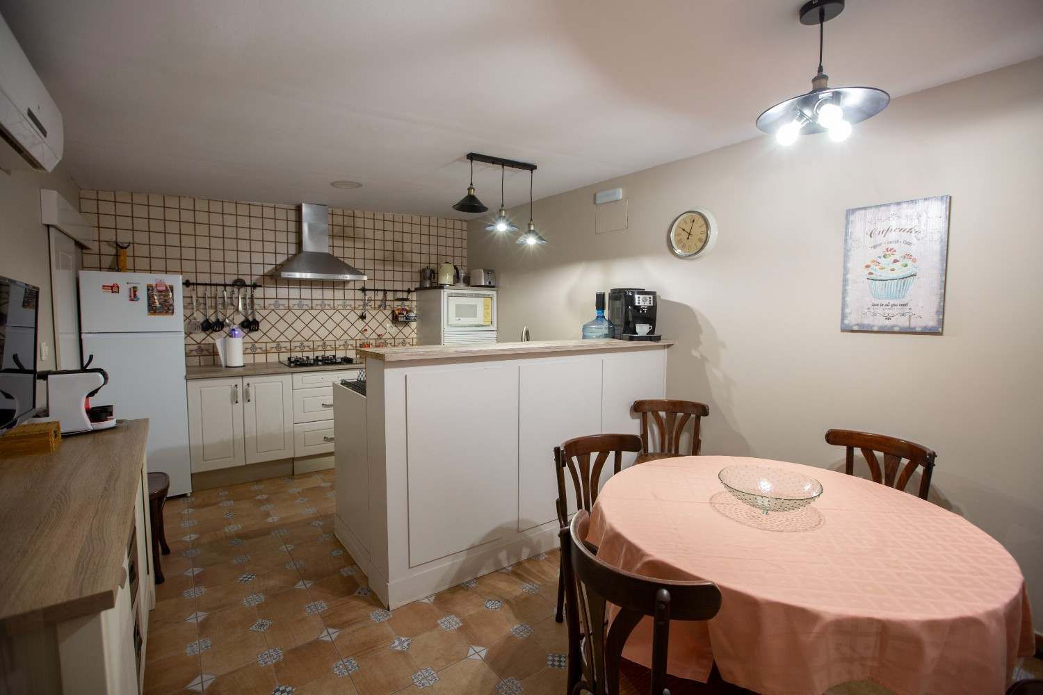 Cocina y comedor con armarios blancos, salpicadero de azulejos, mesa redonda con mantel rosa y sillas.