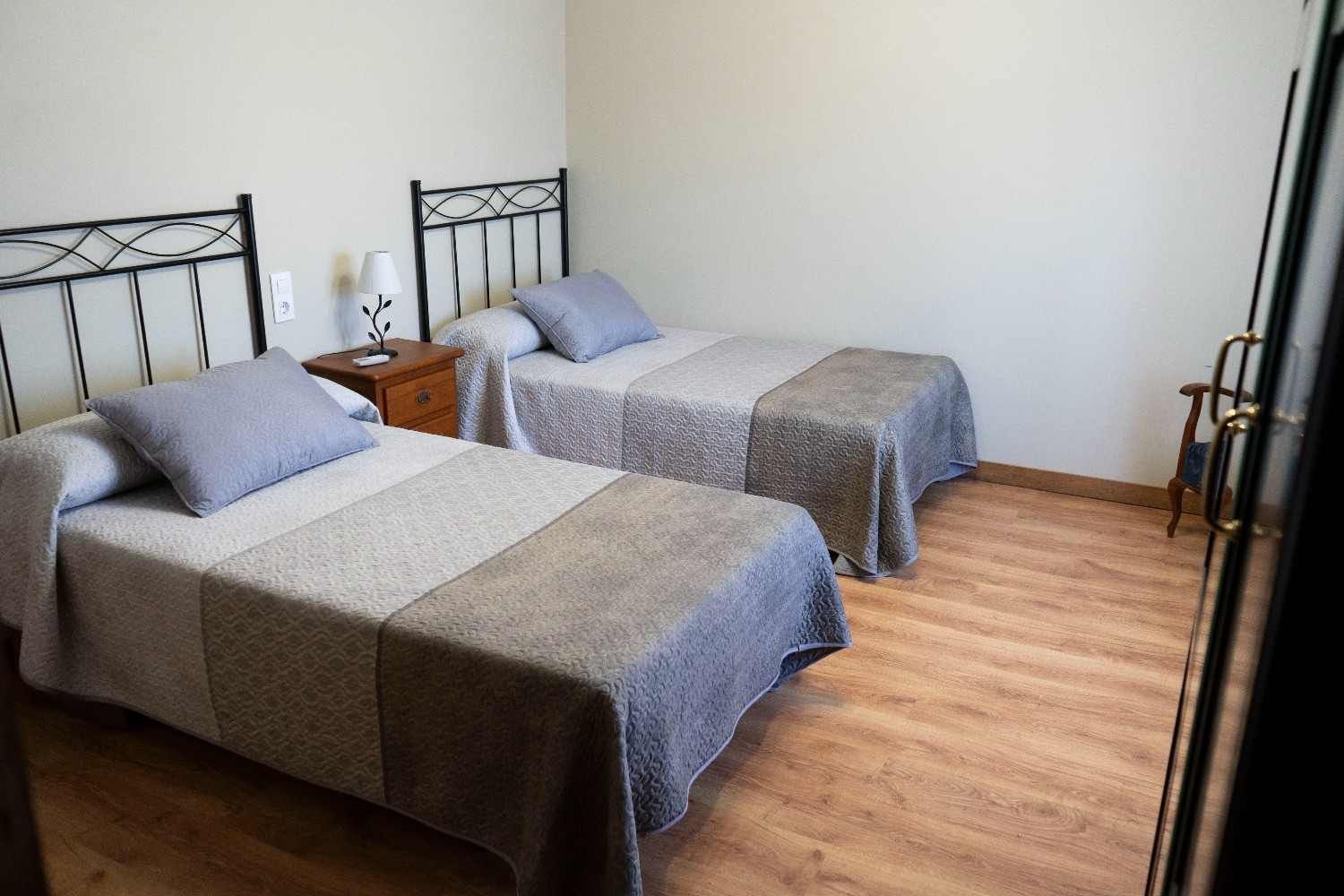 Dos camas individuales con ropa de cama gris en una habitación sencilla con suelo de madera.