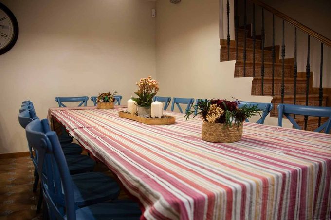 Comedor rústico con mesa de madera, bancos, plantas en macetas y luz solar que entra por dos ventanas.