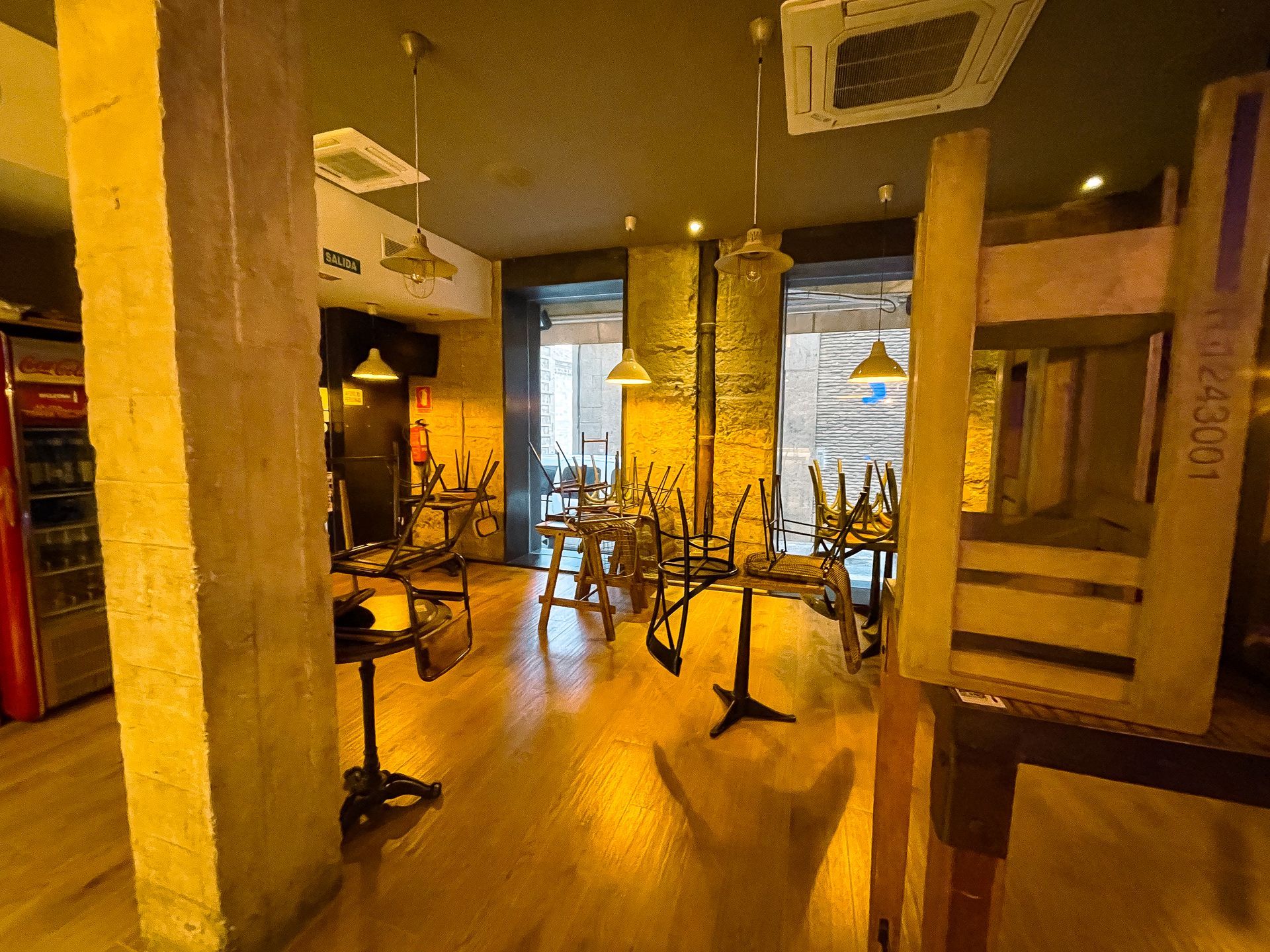 Interior de un restaurante con suelos y columnas de madera. Sillas apiladas sobre mesas; puerta abierta.