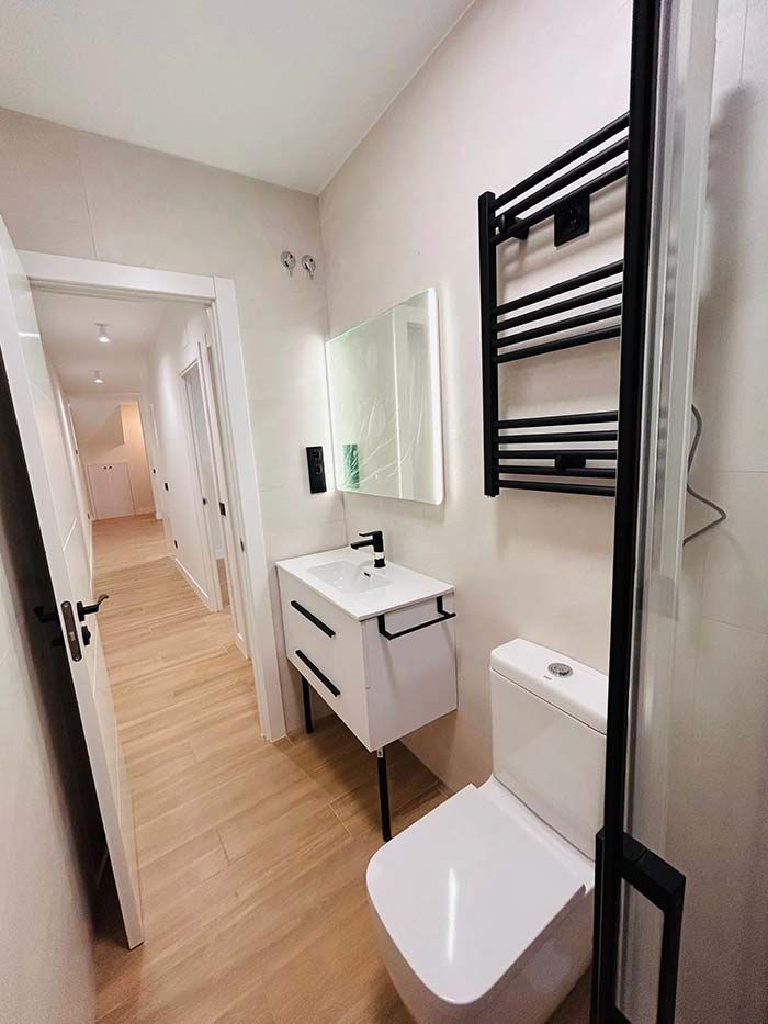 Baño moderno con inodoro blanco, tocador y calentador de toallas negro; la puerta conduce al pasillo.