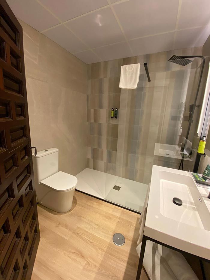 Baño con inodoro, ducha y lavabo. Tonos beige y madera. La ducha incluye toallero y grifería negra.