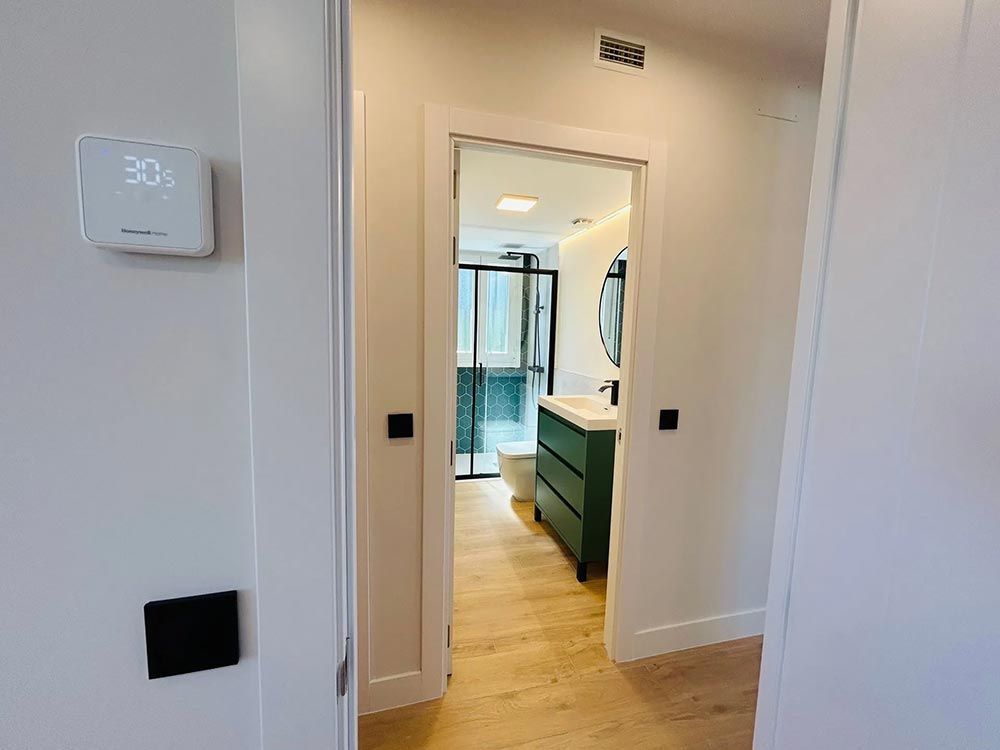 Puerta que da al baño con tocador verde y espejo redondo. Paredes blancas, suelo de madera. El termostato marca 30 grados.