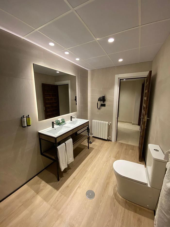 Moderno baño de hotel con lavabos dobles, inodoro y puerta abierta. Suelo de imitación madera clara y paredes beige.