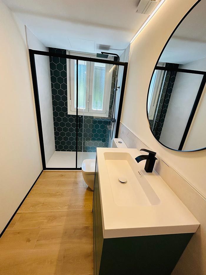 Baño moderno con tocador verde, ducha con marco negro, piso con apariencia de madera y espejo redondo.