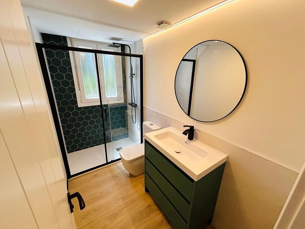 Baño moderno con tocador verde, espejo redondo y marco de ducha negro.