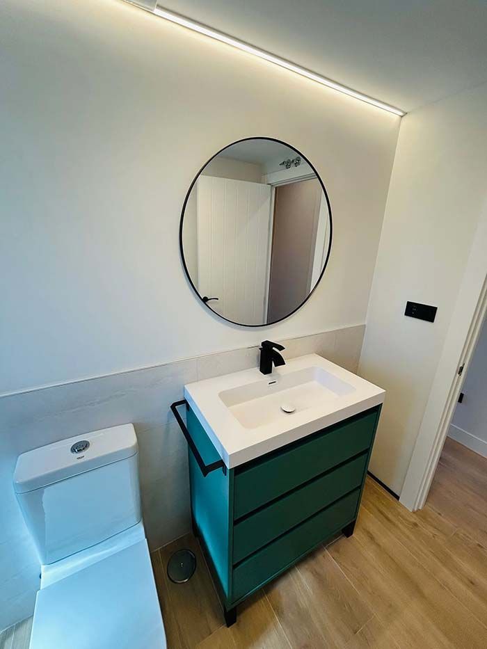 Baño con tocador verde azulado, espejo redondo negro, inodoro y accesorios modernos.