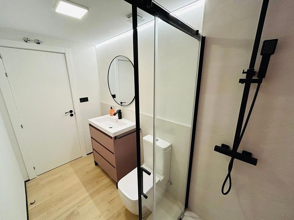 Baño moderno con tocador ruborizado, espejo redondo, ducha de vidrio y accesorios negros.